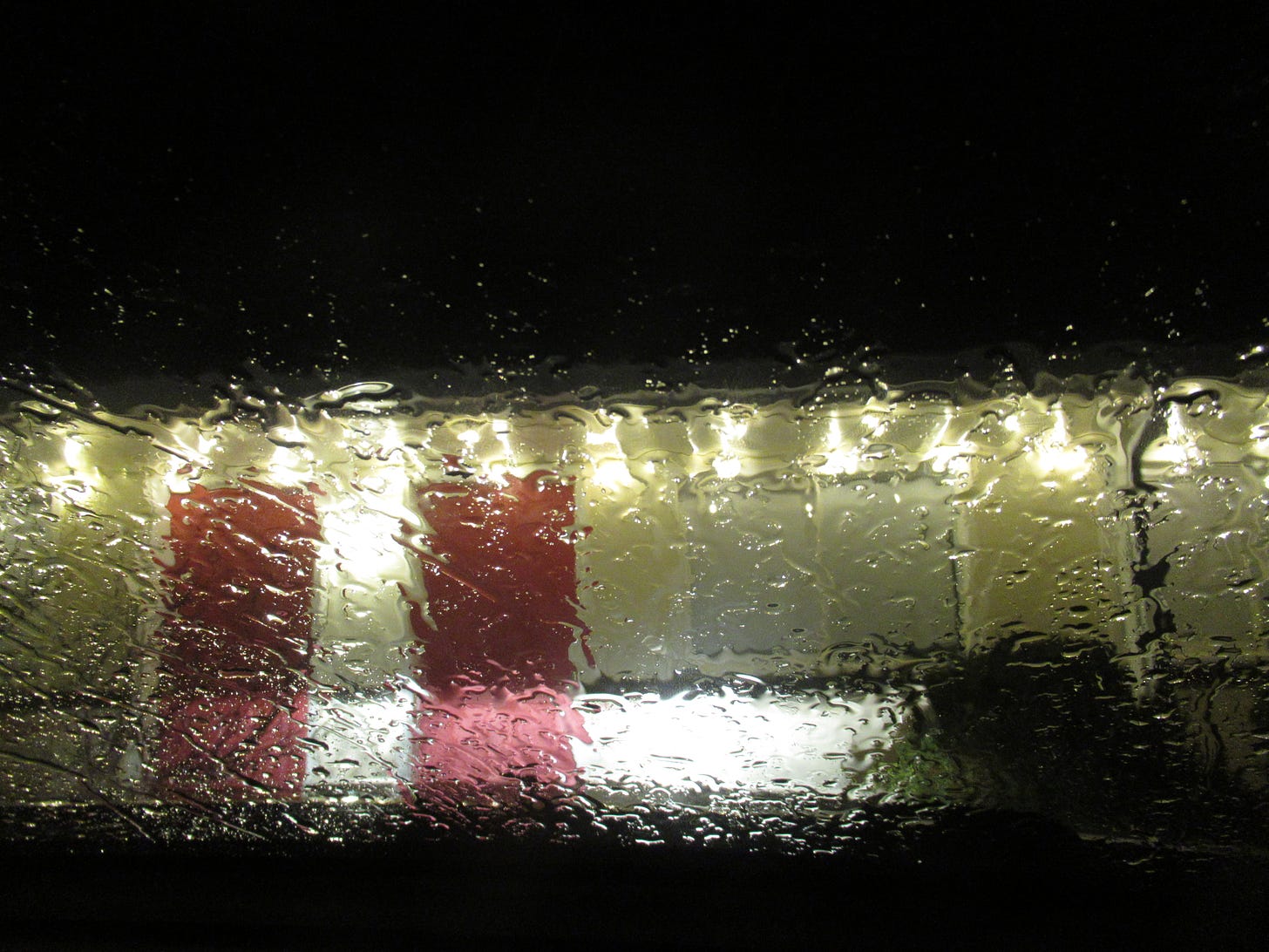 Facade of a vintage motor lodge viewd through the rain streaked windshield of a vehicle in front of it. The doors are bight red and there are blurry lights lining it