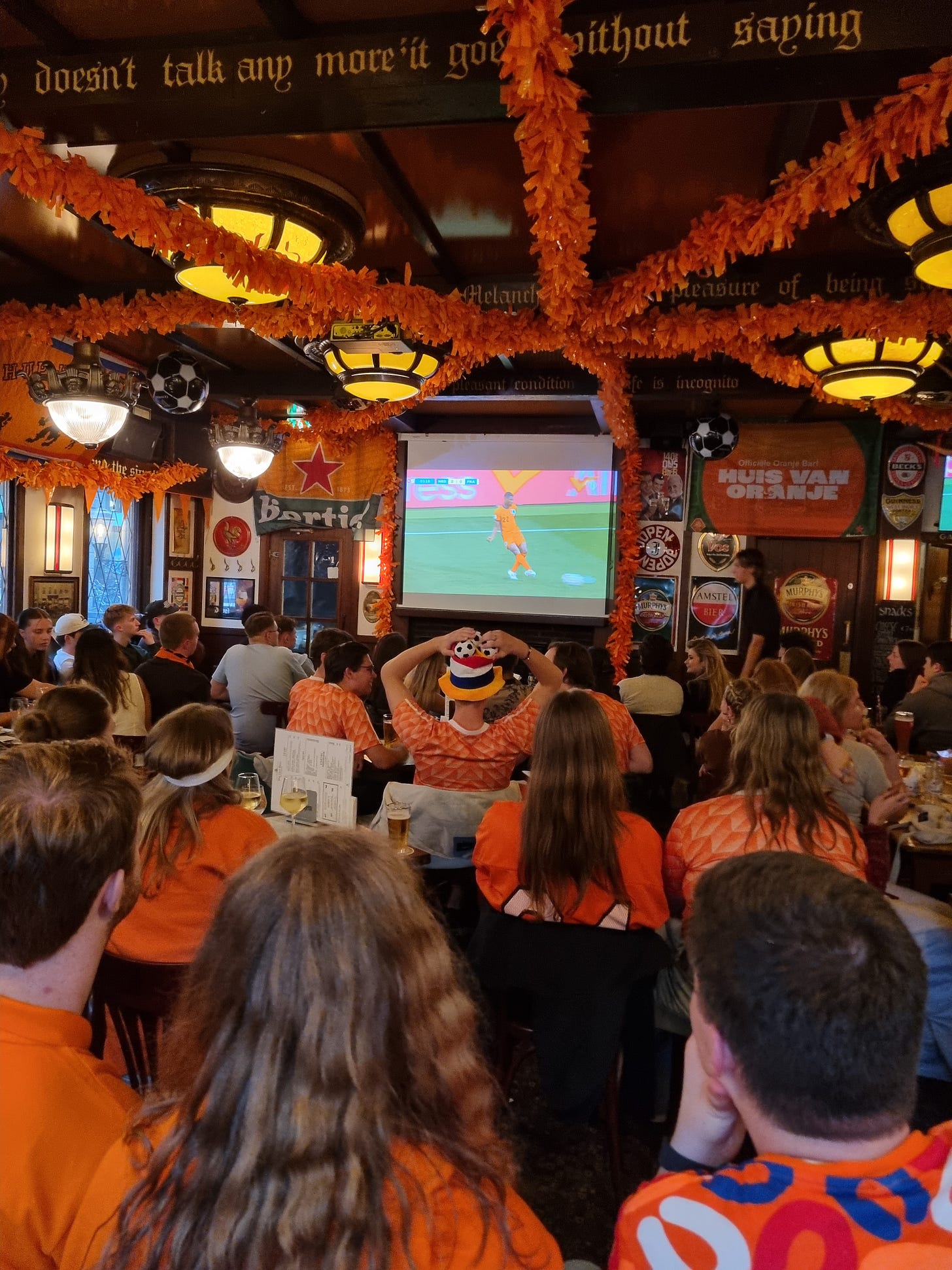 Photo from inside a pub, decorated with orange streamers. People sitting in front are wearing orange and facing a screen showing a game of soccer.