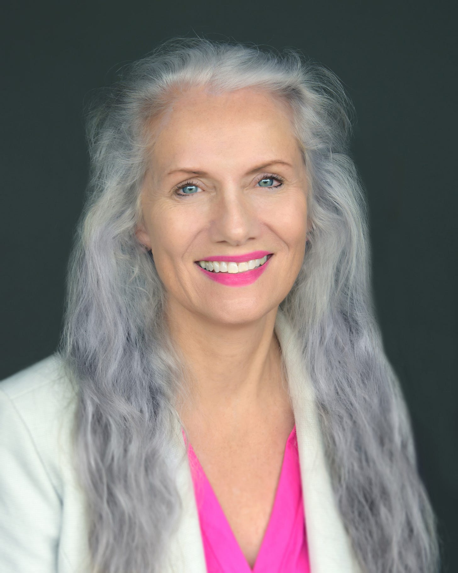 Woman with long gray hair, blue eyes, and bright pink lipstick smiles at camera