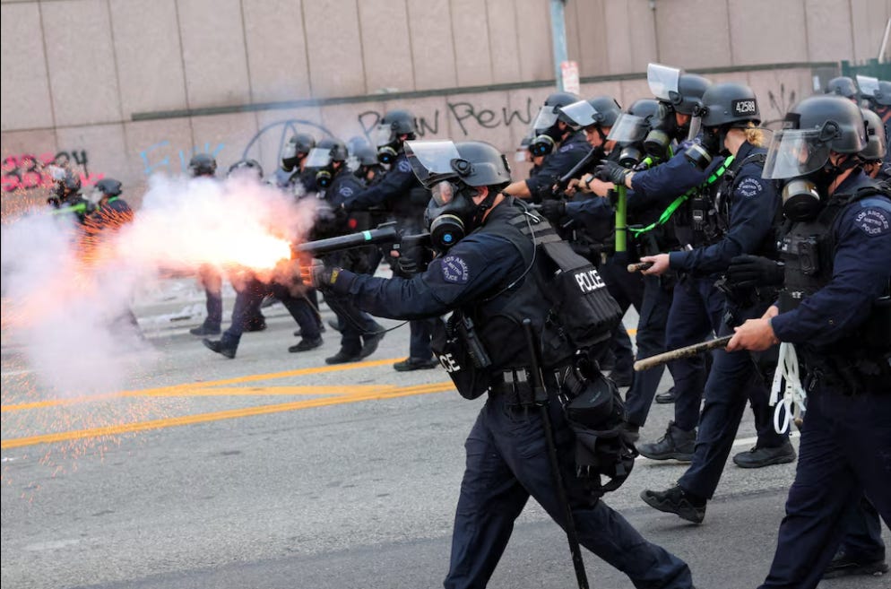 A line of armed forces wearing Police insignia advance in down an urban street. A figure center front fires a weapon in fiery hues.