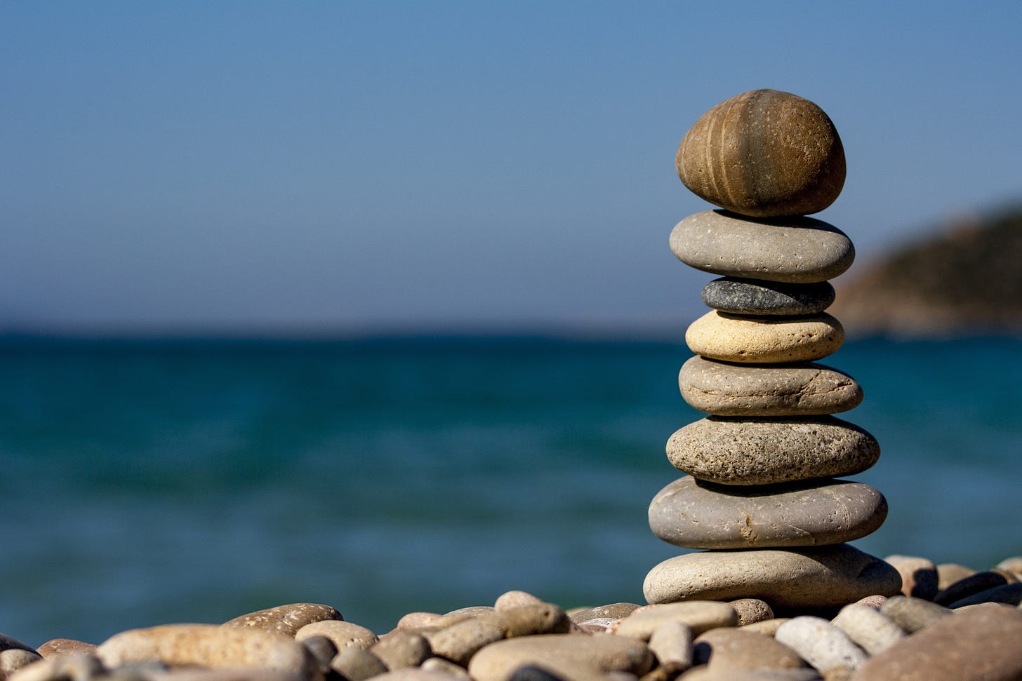 an image of 8 stones balanced on top of each other in a stack on the shore of a lake or ocean with green waters an d a blue sky blurred in the background