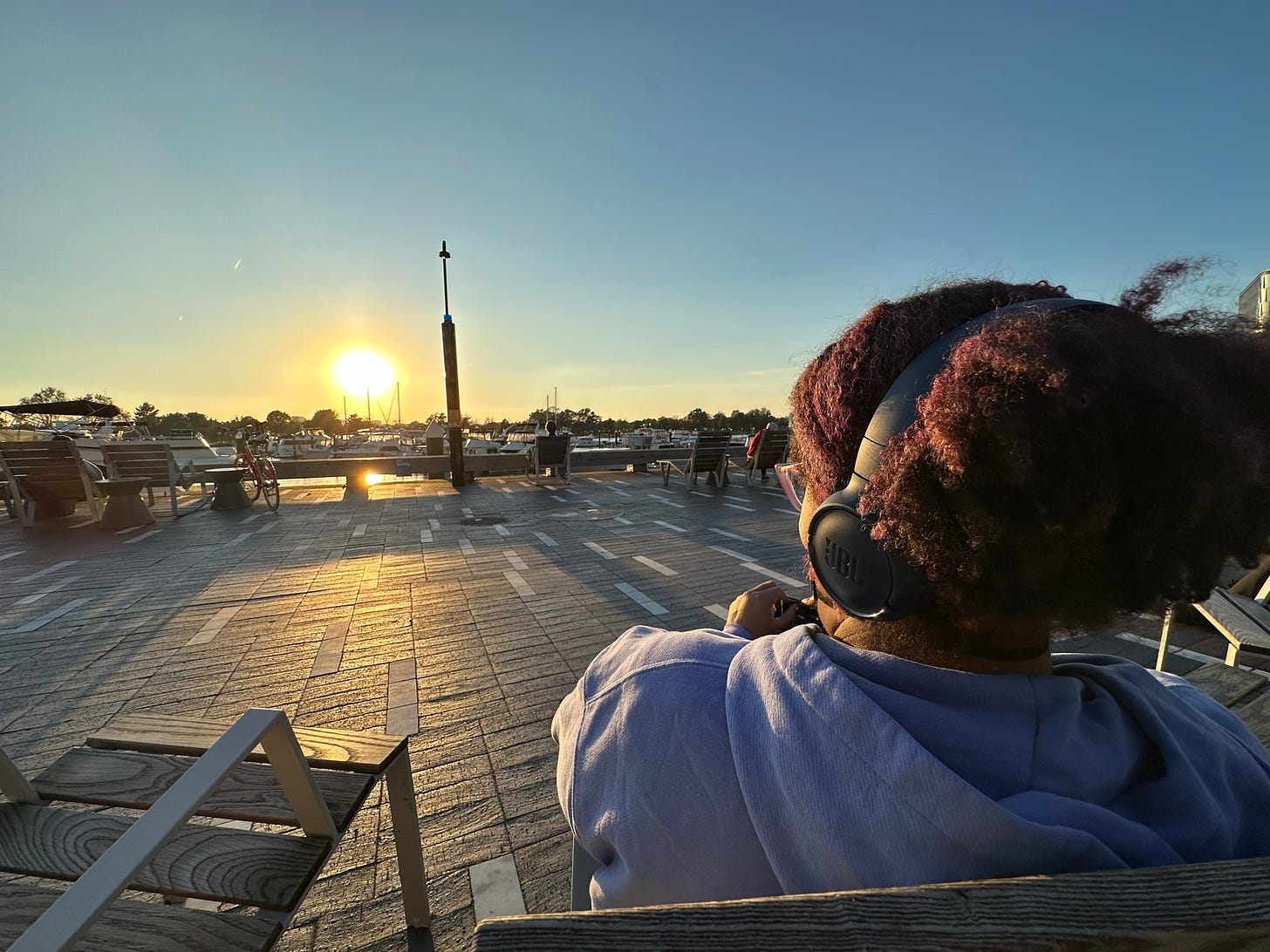 Kristen is facing the sunset  at the District Wharf, sitting in aa metal lounge chair, in a lavender hoodie and with blue headphones on her black and fusha hair