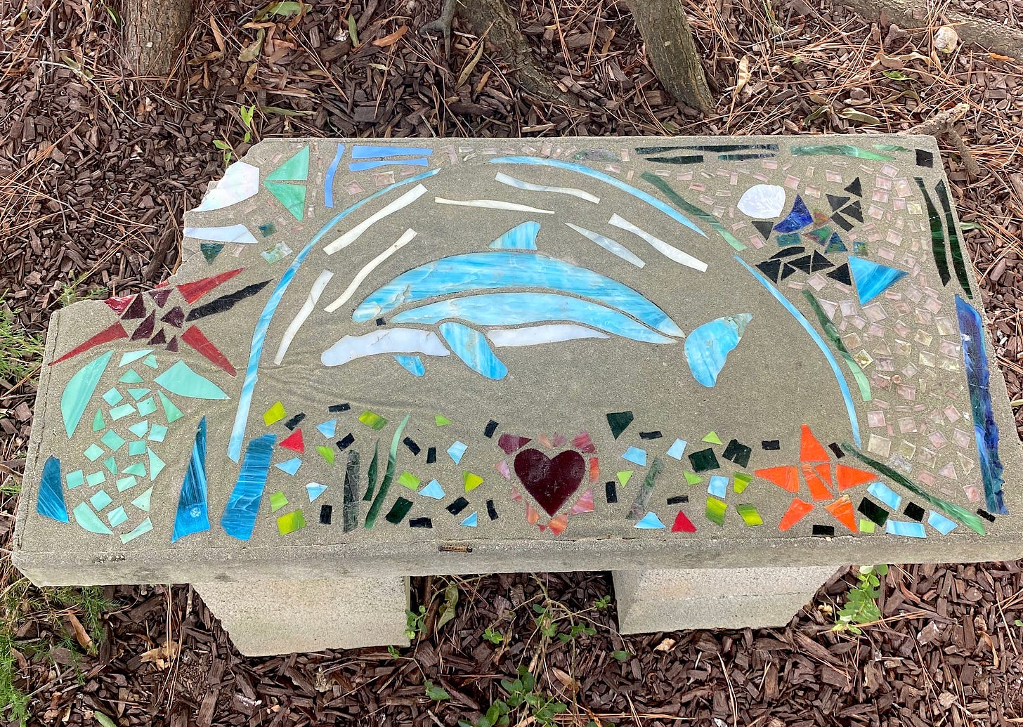 A grey concrete bench decorated with stained glasses images including a blue and white dolphin, sea plants, and a heart. The top left corner is broken off.