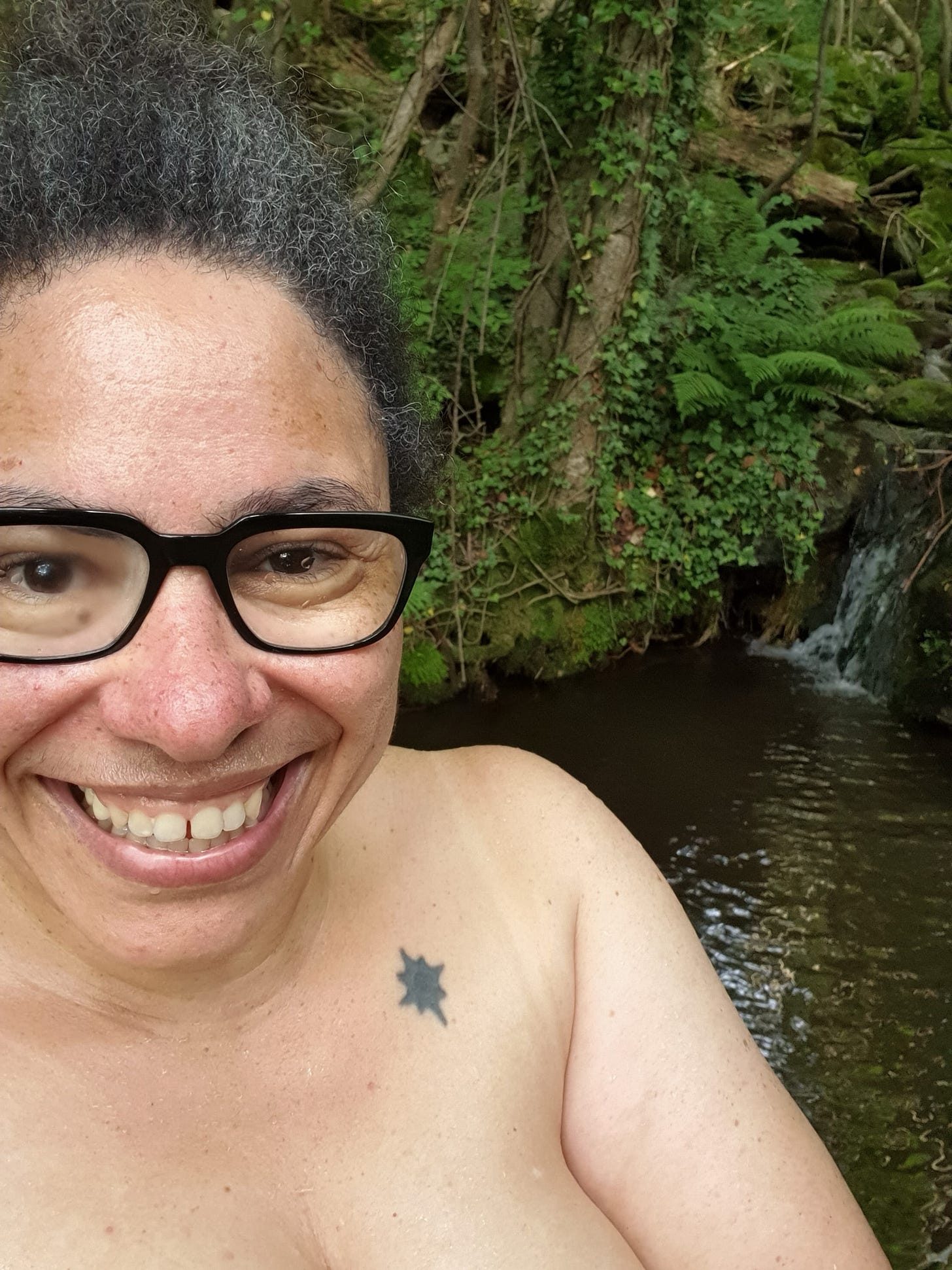 Photo of a woman in black-rimmed glasses in front of a natural pool. She is naked but you can only see from her cleavage upwards.