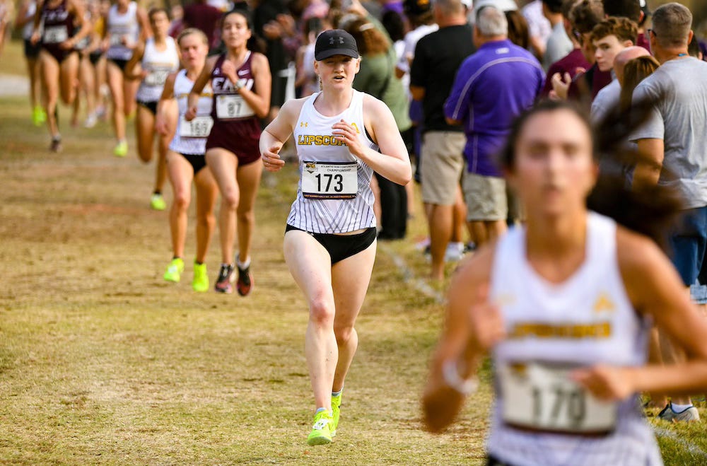 Liza Corso runs in a college cross country race, wearing a white Lipscomb uniform and a black running hat.