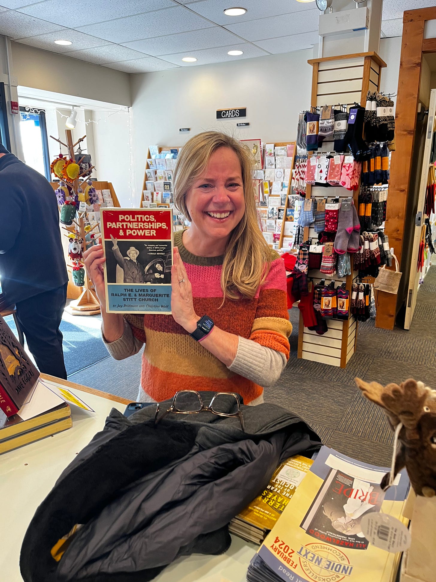 Memoir Coach Christine Wolf holds a copy of her debut book, Politics, Partnerships, & Power: The Lives of Ralph E. and Marguerite Stitt Church in the independent bookstore The Book Stall in Winnetka, Illinois. Wolf is wearing a striped sweater and crying. She's standing in front of a rack of socks and greeting cards.