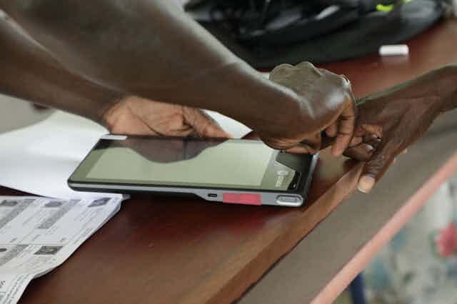 A man assisting another to place a finger on an electronic device that captures biometric details.