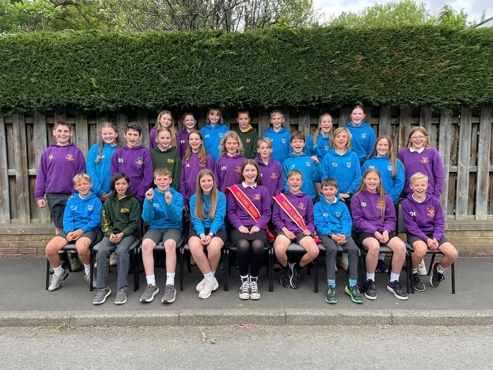 Three rows of P7 pupils from Peebles primary schools are seated for a formal picture to announce this year's Beltane Court.  They are smiling and dressed in the bright blue, purple and green of the town's primary school colours and Annie and Evan are wearing sashes that say Beltane Queen and First Courtier. 