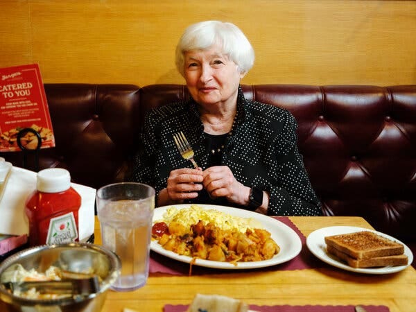 Janet L. Yellen, the Treasury secretary, holds a fork as she sits at a table that has toast, a plate of eggs and home fries, a glass of water and a bottle of ketchup. Next to her is a menu that reads “catered to you.”