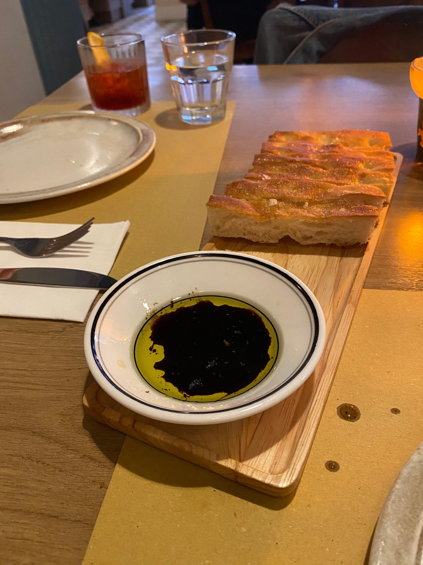 A wooden board with olive oil and balsamic in a white dish a blue stripe around the rip, and focaccia at the other end. The table is set with plates and cutlery, and in the background is a negroni in a hi-ball glass.