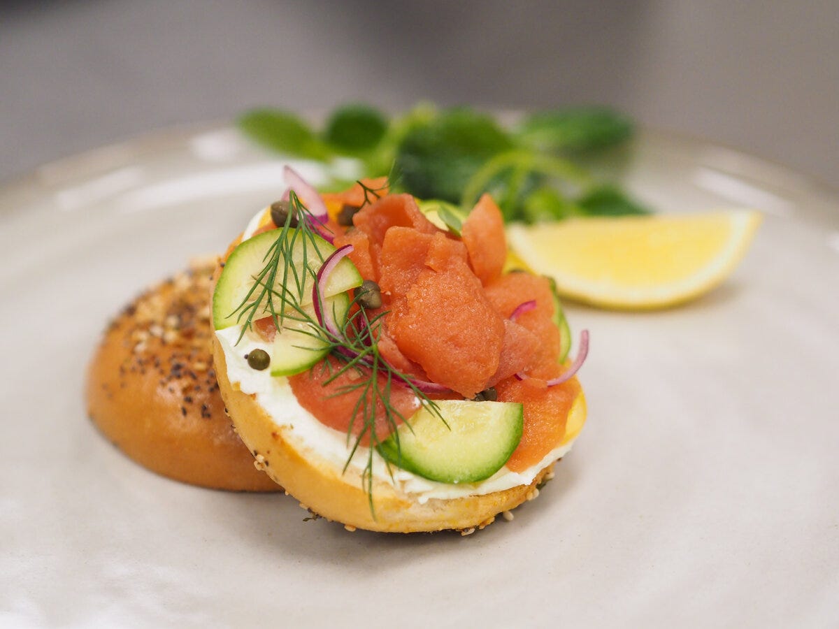 An everything bagel with cream cheese, cucumbers, capers, slices of cultivated lox, and sprigs of dill on a white plate