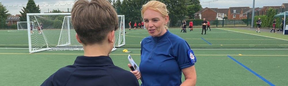 Female Police Community Support Officer with a young footballer with football pitch and goal behind them.