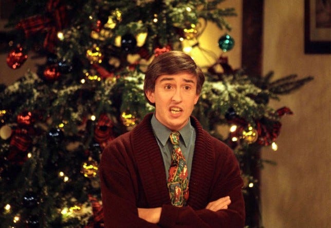 Steve Coogan as Alan Partridge in front of a christmas dress wearing a cardigan and jazzy tie
