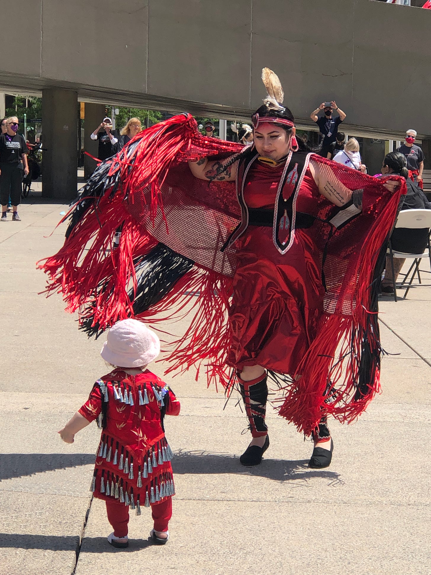 Dancer in Indigenous regalia is dancing beside a child, who is watching dancer. The child is also wearing Indigenous regalia.
