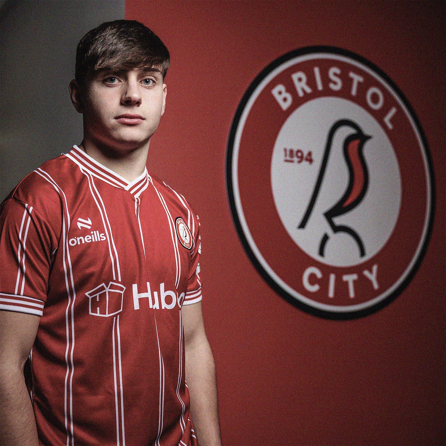 A photo of Josh Stokes wearing a Bristol City shirt while stood in front of a red wall which has the Bristol City crest on it. He's looking directly at the camera. Photo courtesy of Bristol City.
