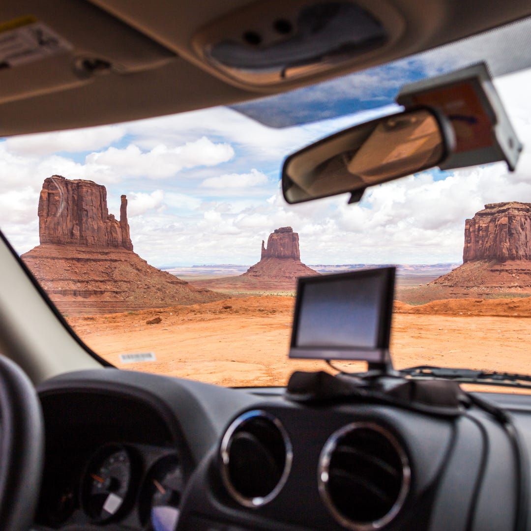 view of Monument Valley, a desert landscape punctuated by odd red rock spires and towers, as seen from inside a car with a GPS mounted on the dash