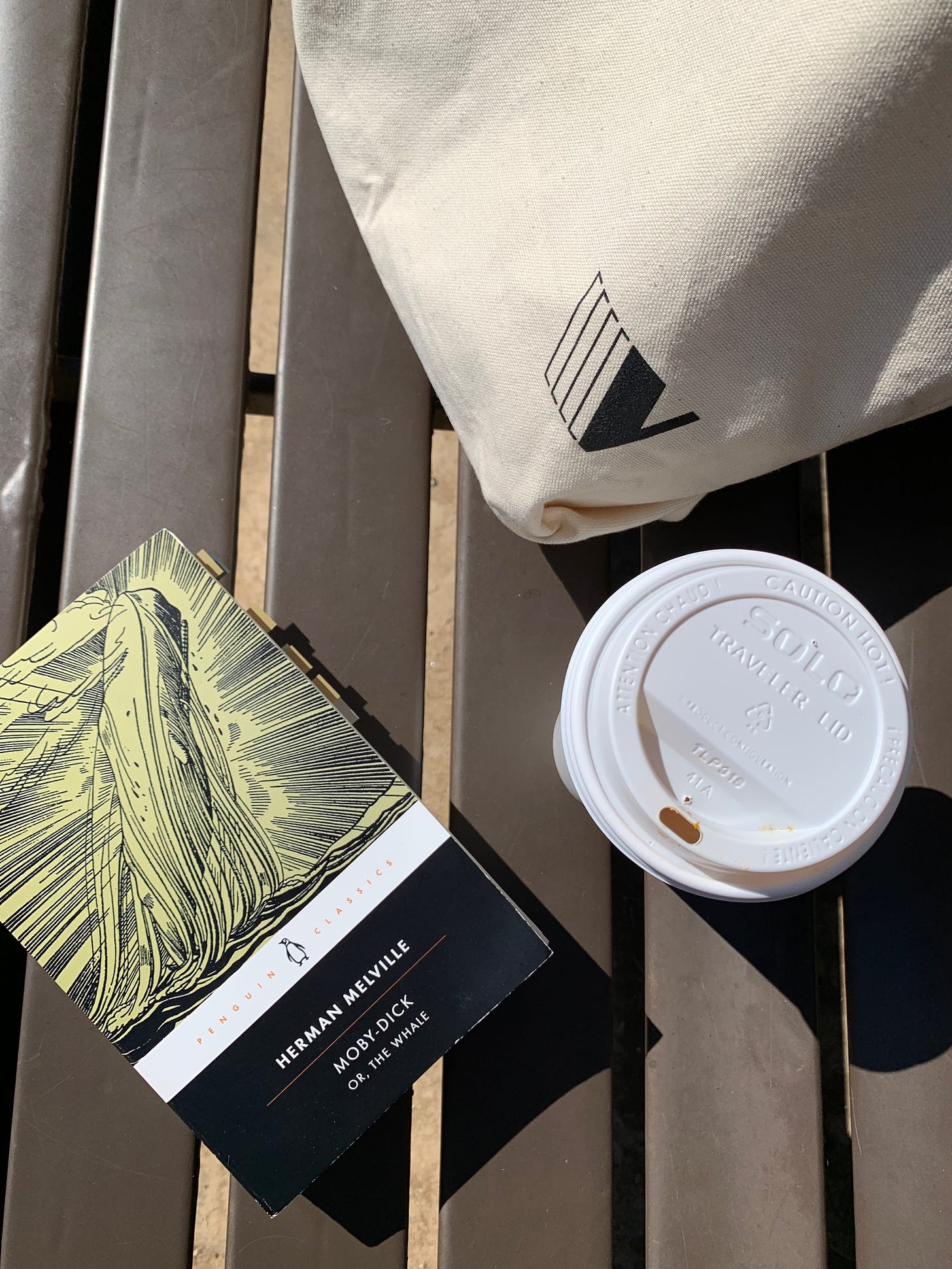 a copy of Moby Dick sits on a bench, next to a coffee cup and a tote bag.