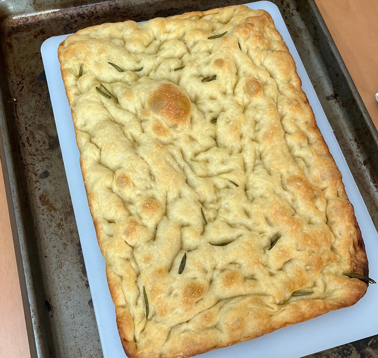 a rectangular loaf of focaccia bread, golden and crispy around the edges, with lots of little craters and folds throughout and some rosemary. The bread is on a white cutting board, on top of a baking sheet.