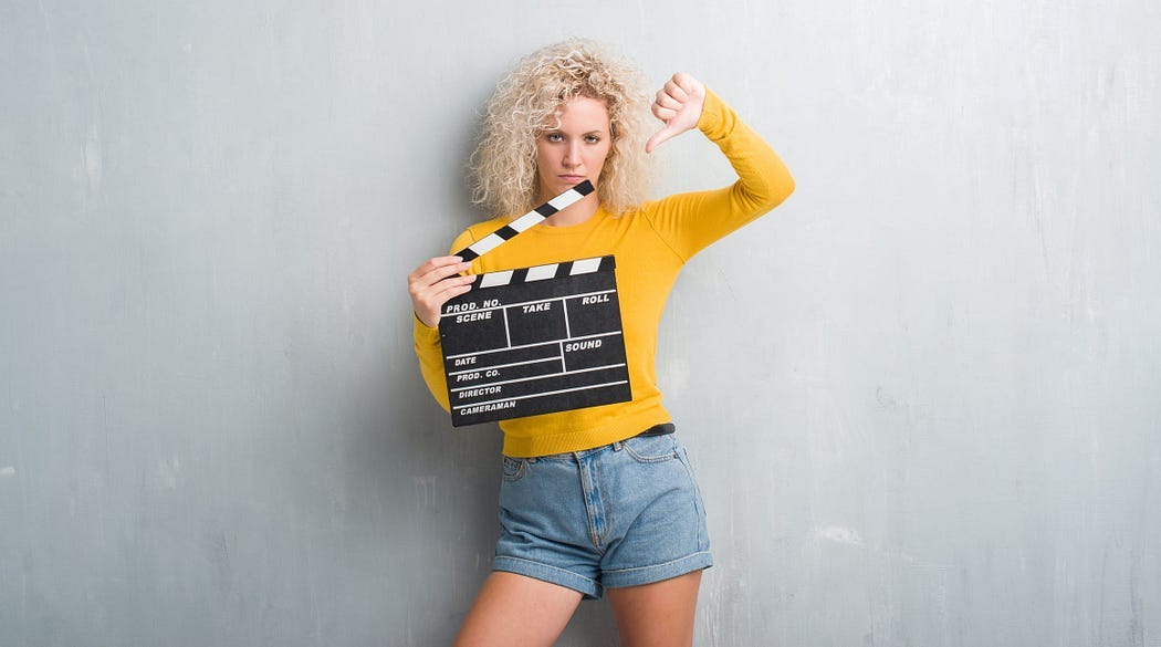 Woman (presumably and actor) in denim shorts and cropped sweater is standing in front of a blank background. She is holding a “clapper” in one hand while making a thumbs down gesture with her other hand.