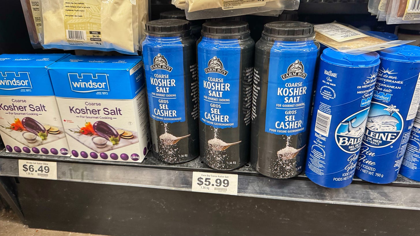 A shelf at a Farm Boy grocery store holds tall, round plastic containers of Farm Boy kosher salt next to boxes of Windsor kosher salt and La Baleine French sea salt