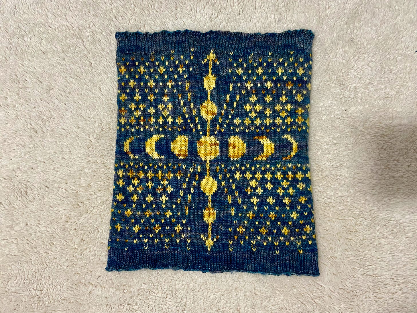 The Ixchel Cowl, which has a variegated blue background and variegated yellow stars, moons, and an arrow, laying on an off-white blanket.