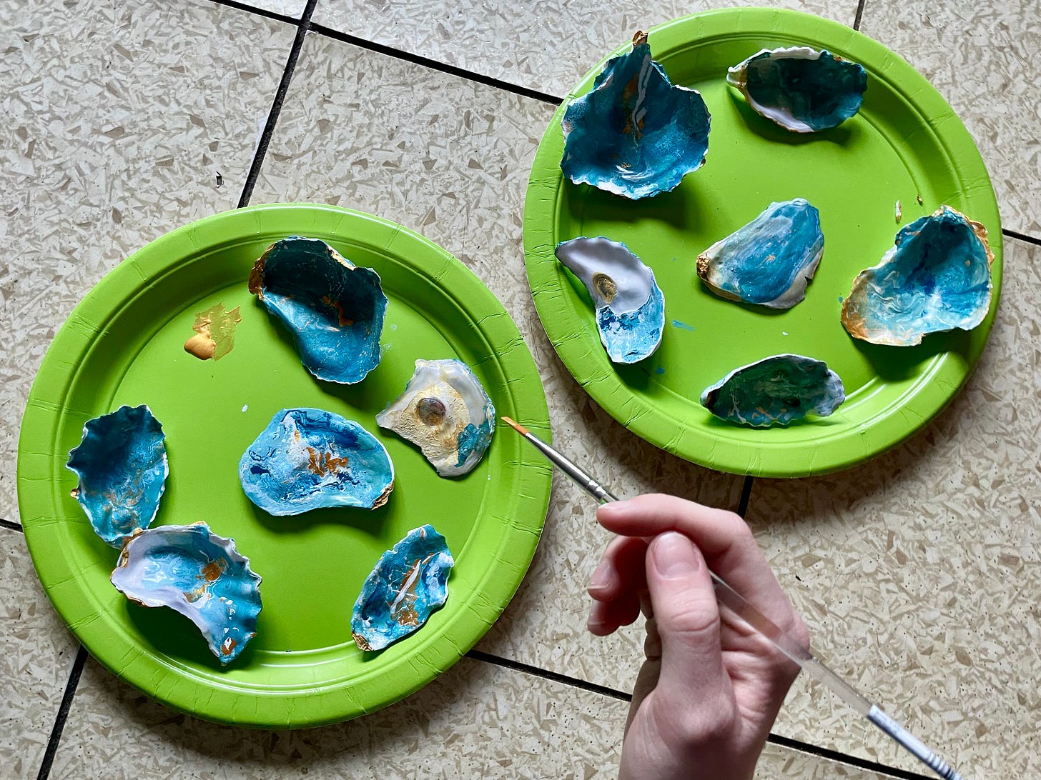 A photograph of two green plates, covered in hand-painted oyster shells. There is a hand holding a paint brush with some gold paint on the tip.