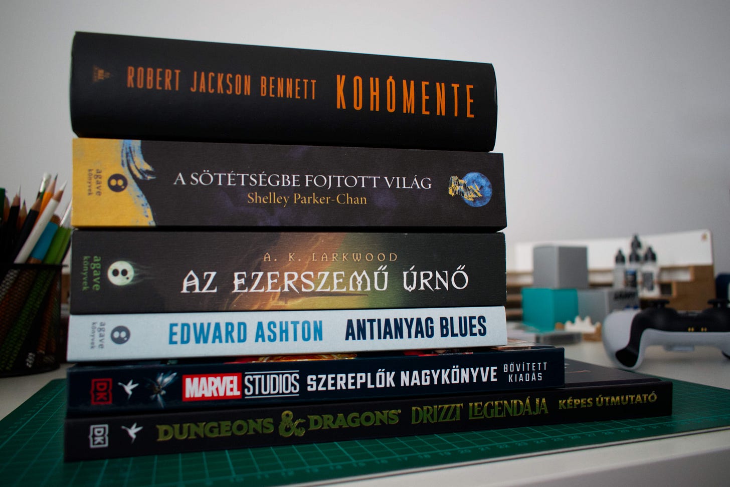 A stack of books laying on their sides on a white desk with pencils and paints in the background.