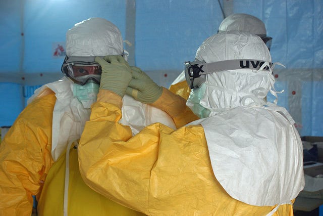 Un operatore sanitario aiuta Jordan Tappero, allora direttore della divisione Global Health Protection dei CDC a indossare la maschera protettiva prima di entrare nell'unità di trattamento di Ebola ELWA 3 allestita da Medici Senza Frontiere ad agosto del 2014 a Monrovia in Liberia. Credit: CDC Global / Flickr. Licenza: CC BY 2.0. 