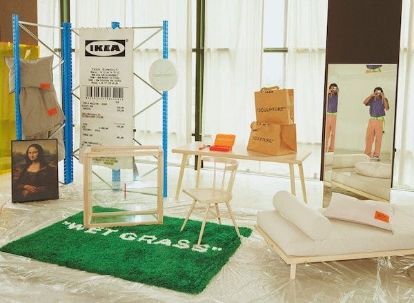 A scene featuring a green rug with print that says “Wet Grass”, a chair, table, bed, and a person taking a photo in a mirror.