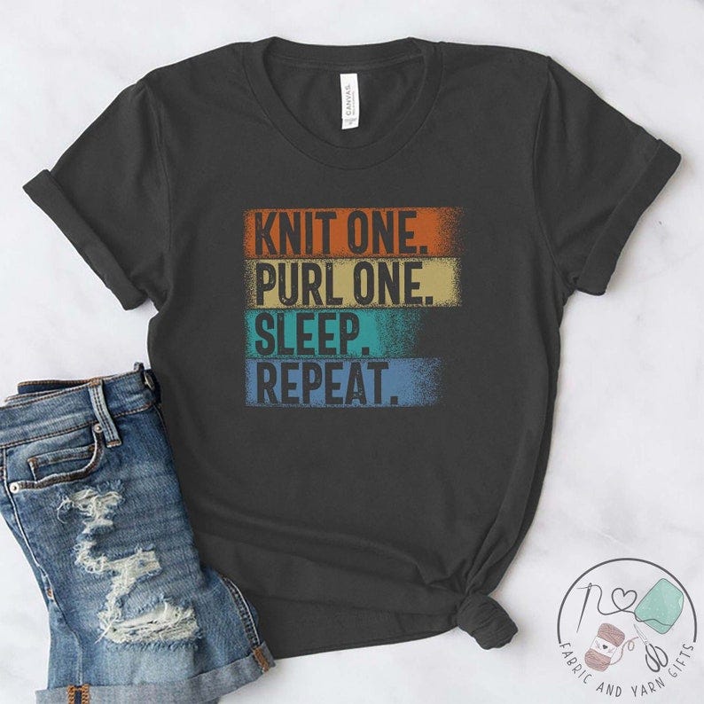 May include: A dark gray t-shirt with the text 'Knit One. Purl One. Sleep. Repeat.' in a vintage style font with colorful stripes behind the text.