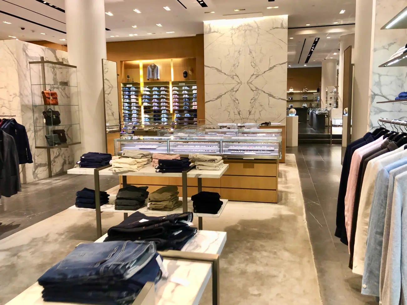 This image depicts the first floor interior of Barneys flagship store on Madison Ave. Neat piles of denim and casual pants are layered on tables with shelves of formal shirting in many colors displayed on a wall in the distance.