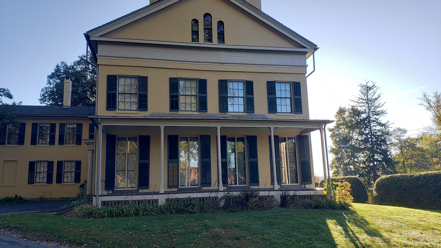 Emily Dickinson's house in the Federal Architecture style. Painted yellow with dark green shutters. A wide porch with four columns. The house has a long addition on the side in the same style. Sitting on a green hill, surrounded by green and gold trees and a clear blue sky. 