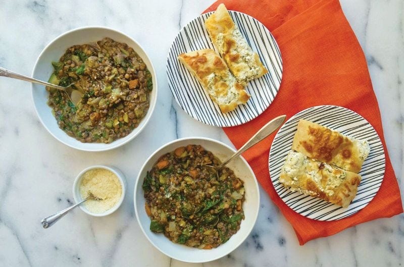 Two bowls of lentil stew sitting on a marble table top with two plates of herb goat cheese focaccia on top of an orange fabric napkin