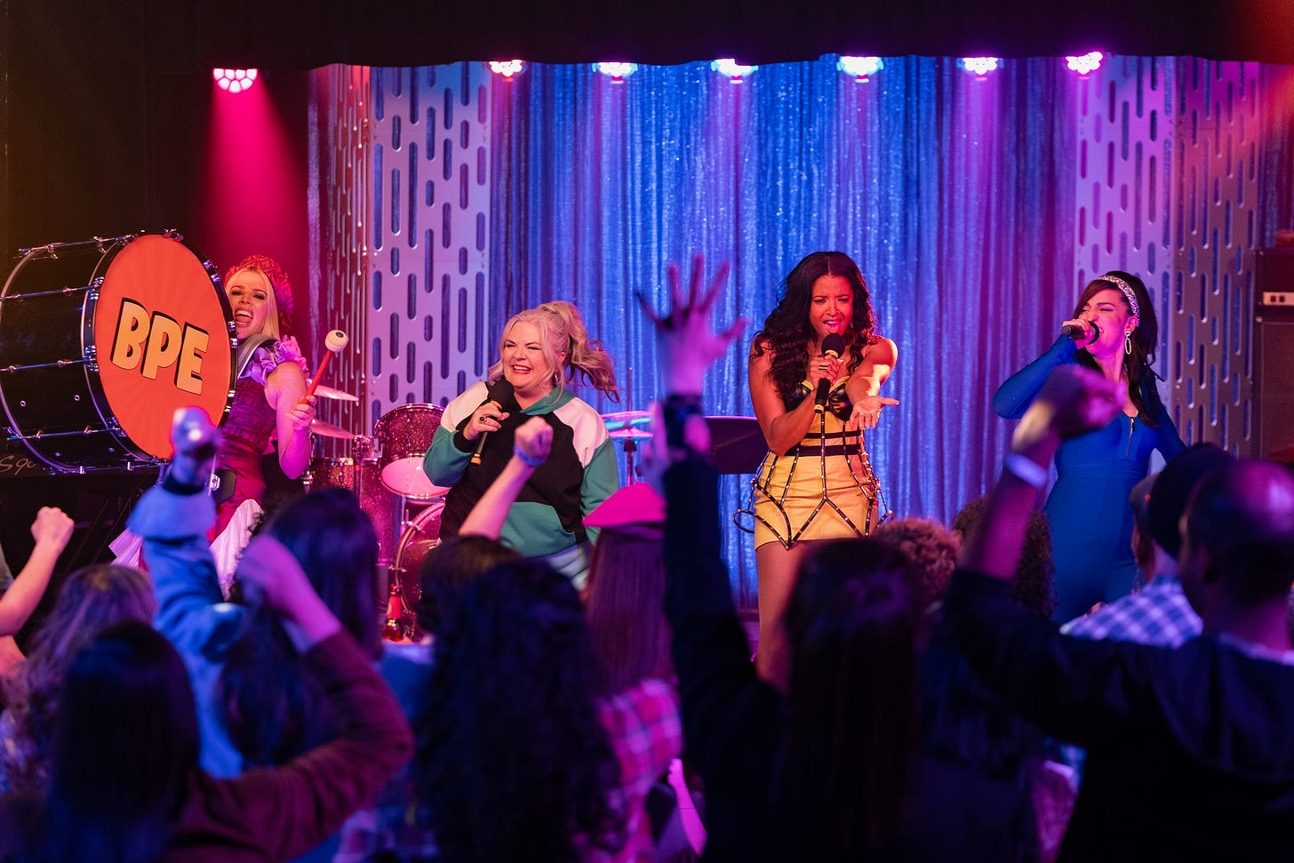 (l-r) Busy Philipps as Summer, Paula Pell as Gloria, Renee Elise Goldsberry as Wickie, and Sara Bareilles as Dawn in GIRLS5EVA. The women are performing a song onstage while Summer is wearing a marching drum with BPE written on it.