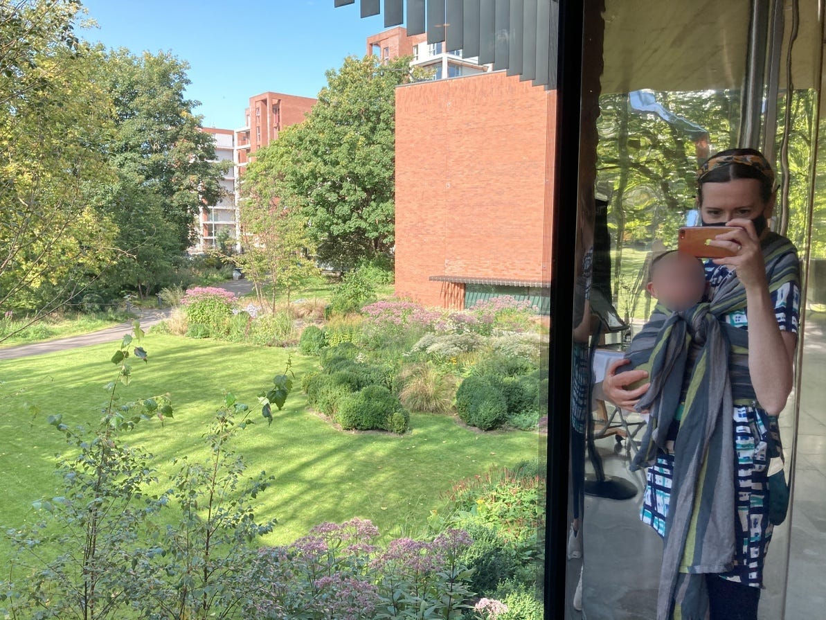 A white woman talking a photo of herself in a mirror. She is carrying a baby in a grey woven wrap. To the left of the image a beautiful garden is depicted