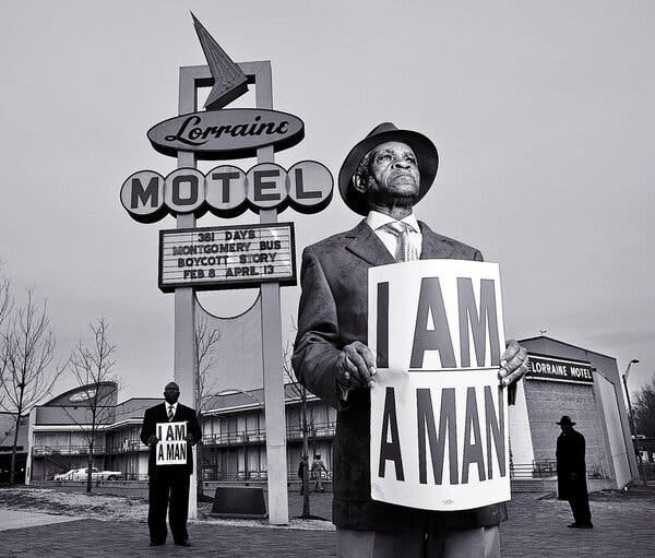 A black and white photograph of Elmore Nickleberry wearing a suit and a hat and holding a sign that says "I AM A MAN." A man standing behind him holds a similar sign. They are in front of a big sign that says "Lorraine Motel."