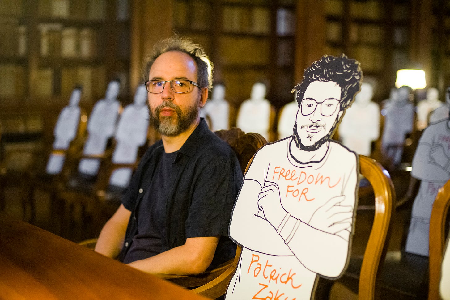A bearded man in short sleeves sits next to a cardboard cartoon cutout of a man