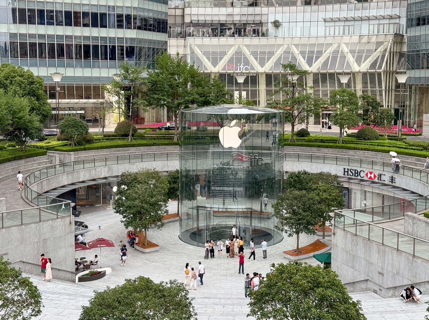 The new glass drum entrance to Apple Pudong and surrounding redesign plaza, pictured from a high vantage point at a distance. Visitors mill about in front of the store.