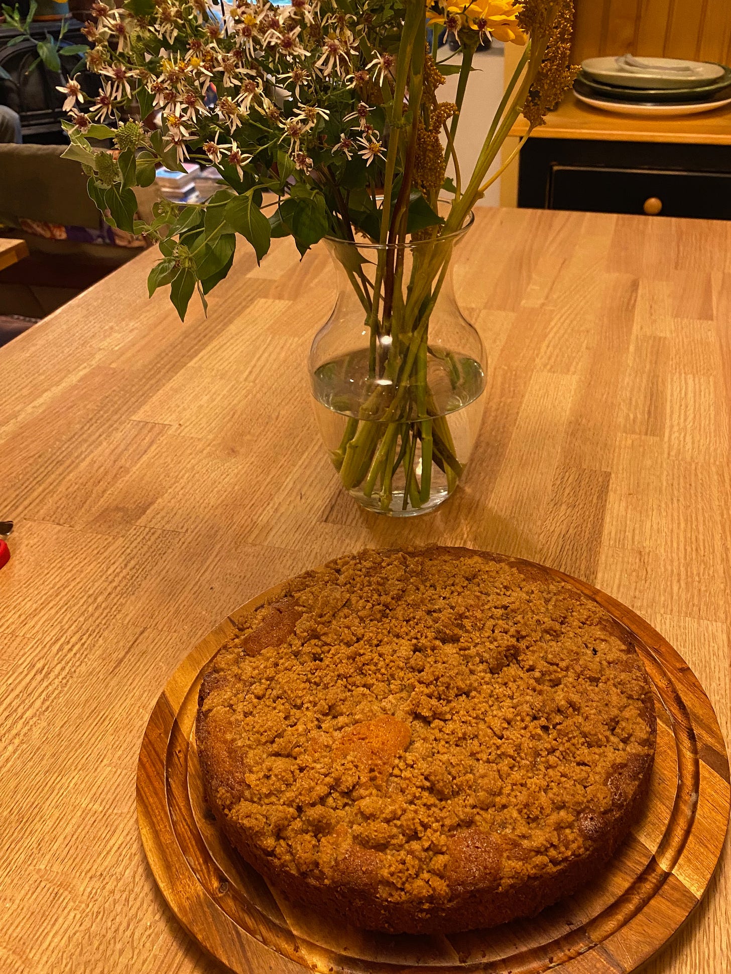 A round cake with a crumb topping sits on a wooden cake board next to a vase of flowers.