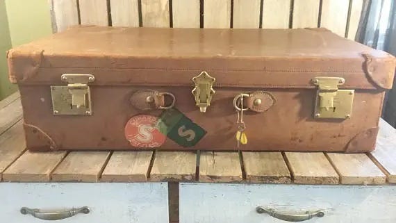 The front of a large brown trunk. There are a couple of stickers on it and keys hanging out of a lock. It has no handle.