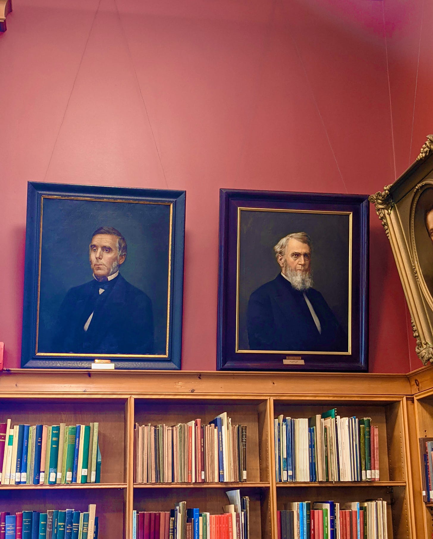 A library wall, painted dark pink with several oil portraits of older, white men from the 1800s, in ornate frames. The paintings hang above several shelves of library books.