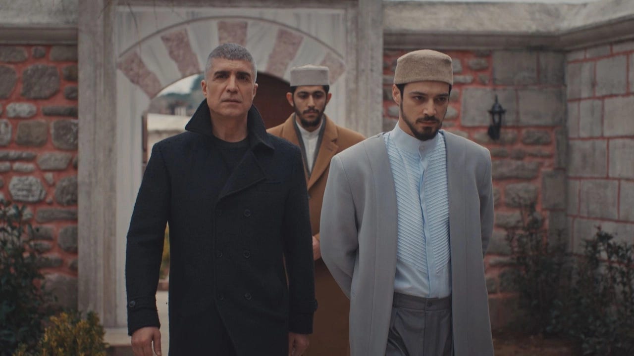 Dört erkek karakter, taş duvarlarla çevrili tarihi bir avluda yürürken görülüyor. Arka planda kemerli bir giriş kapısı ve bahçe detayları bulunuyor.