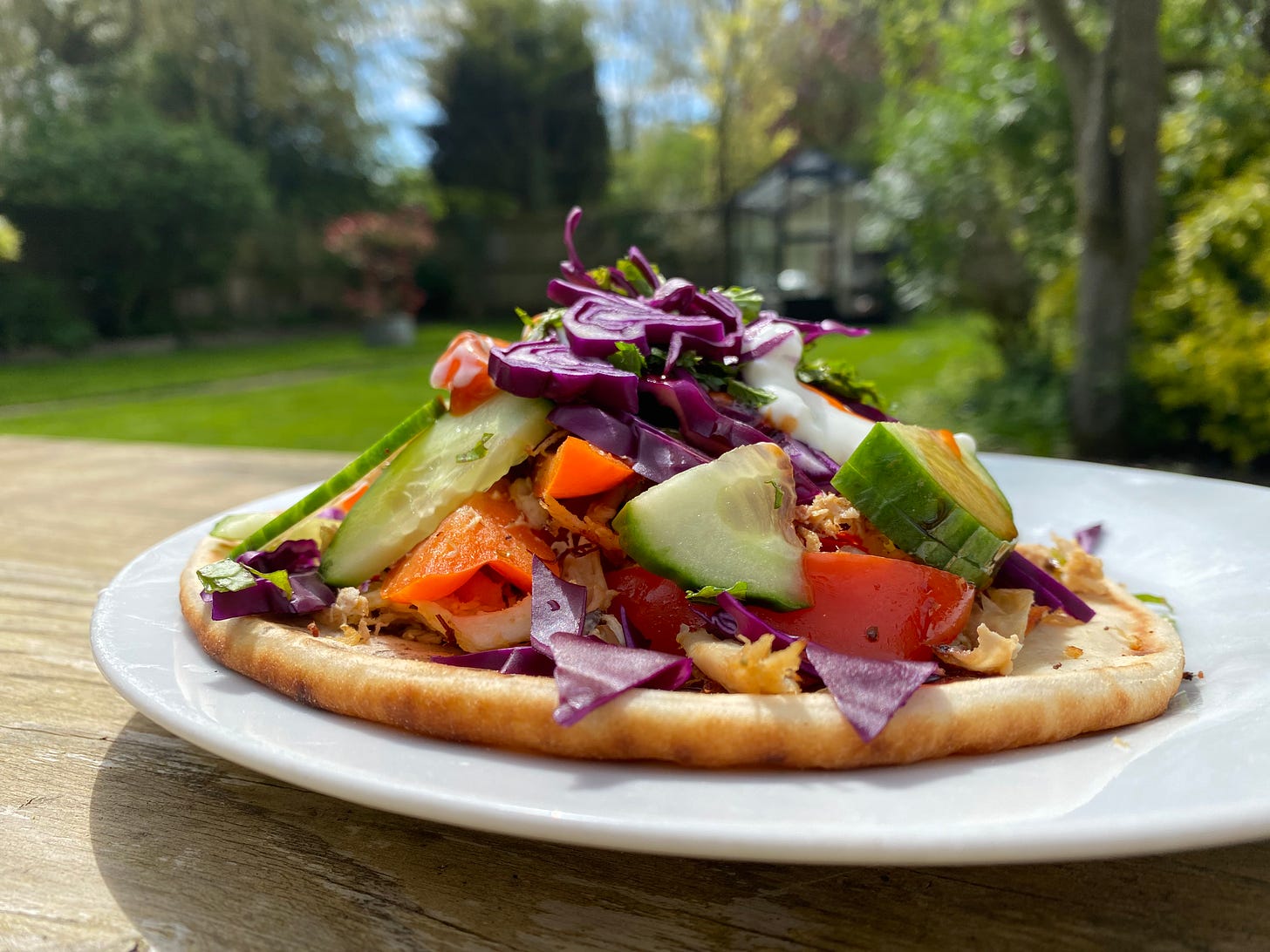 Plate with flatbread topped with cucumber, red cabbage, peppers, and green herbs. The plate is on a table in a garden.