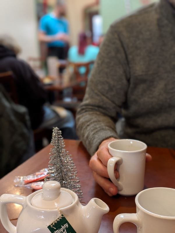 a hand holding a coffee mug on a table that features a small tea pot and another mug, with a waiter talking to patrons in the background