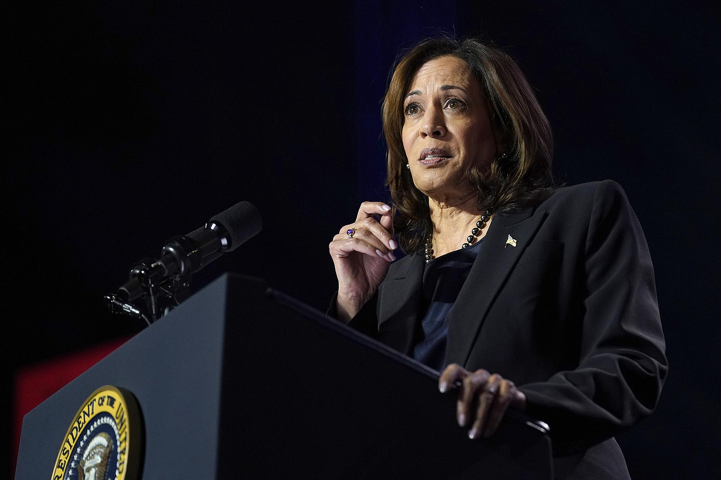 Vice President Kamala Harris, an Indian and Black woman with medium skin tone wearing a black suit, talks while standing in front of a podium. 