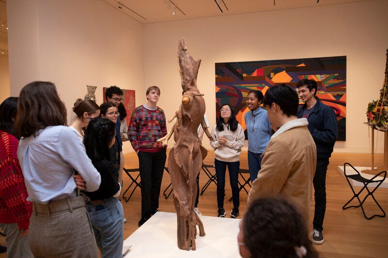 A group of people standing next to a work of sculpture in a gallery. They direct their attention to the sculpture, which resembles a human figure but with elements of tree branches. A colorful artwork hangs on the wall behind them.