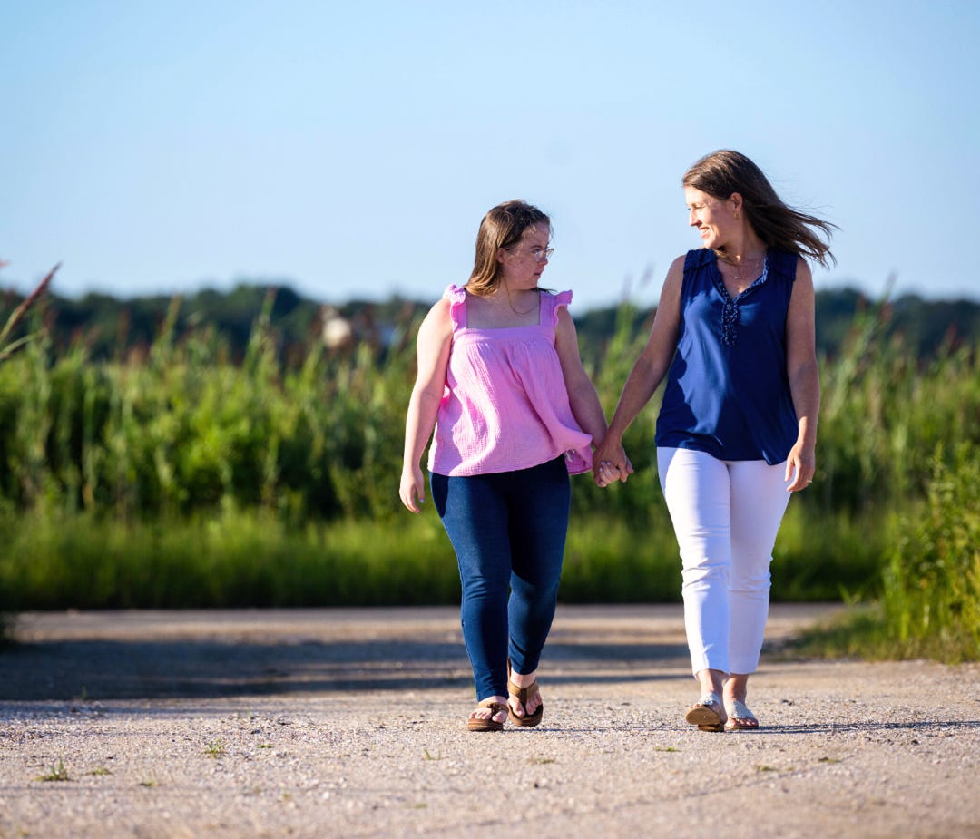 Penny and Amy Julia are walking hand in hand outdoors on a sunny day. Amy Julia is looking down at Penny and smiling warmly as they share a moment. They are walking along a dirt path with green grass and tall plants in the background, under a clear blue sky.