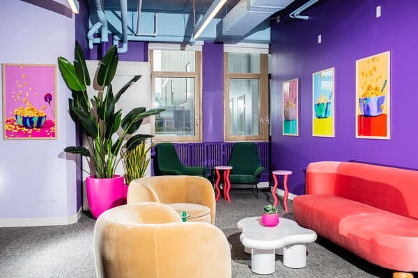 A bright room that has purple walls, framed artwork of cereal bowls, a pink velvet couch, yellow velvet chairs and tables in squiggly shapes.