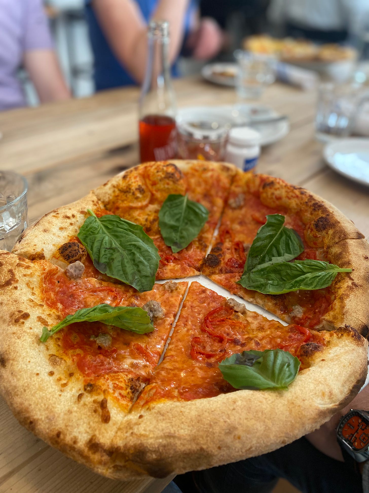 A wood-fired Italian pizza with sausage, peppers, and large basil leaves. In the background is a small bottle half-full of a red bicicletta cocktail, and a glass beside it holds the other half.
