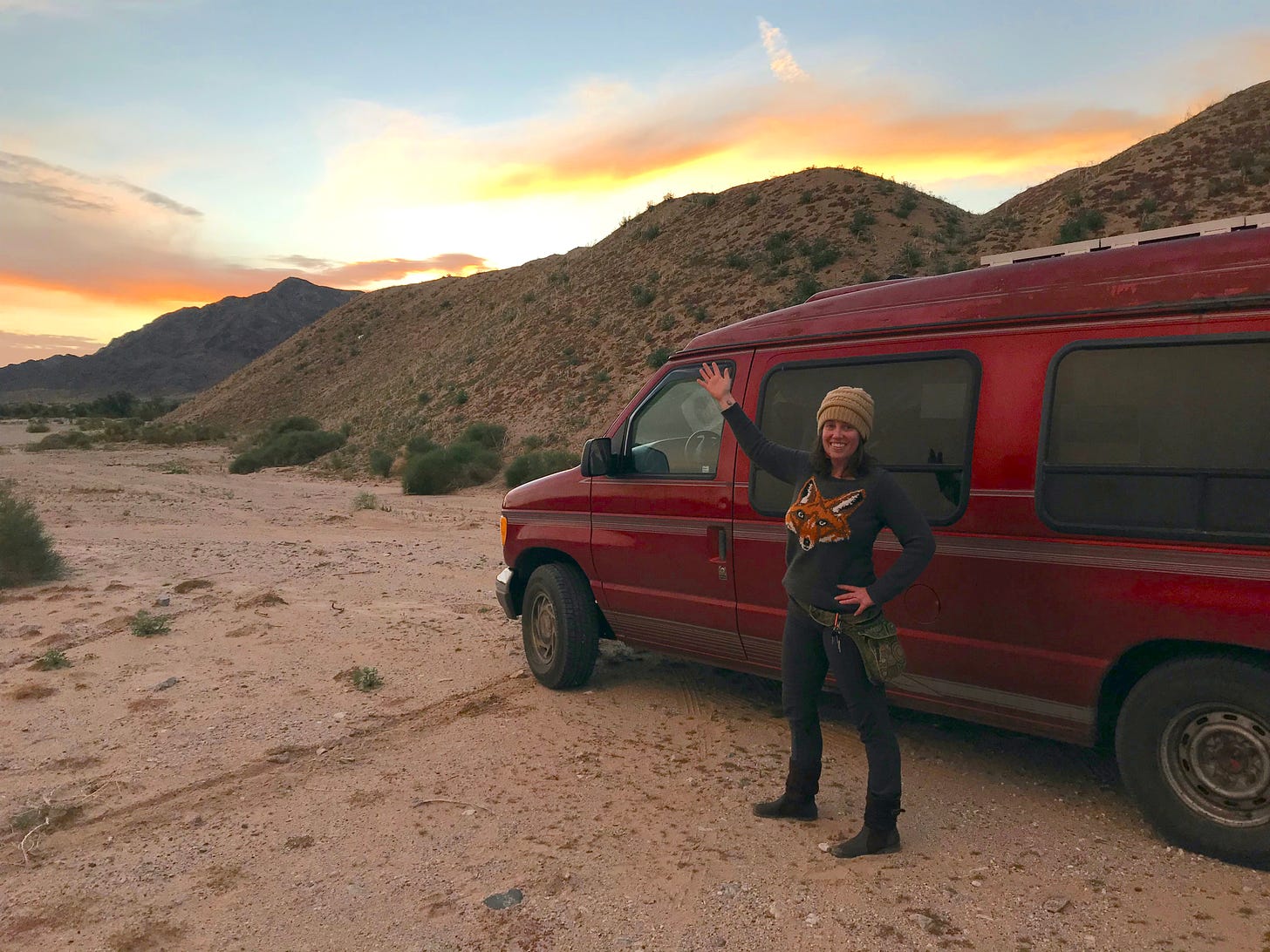 A woman (the author) stands in front of a red van pointing to the sandy hills and sunset behind her